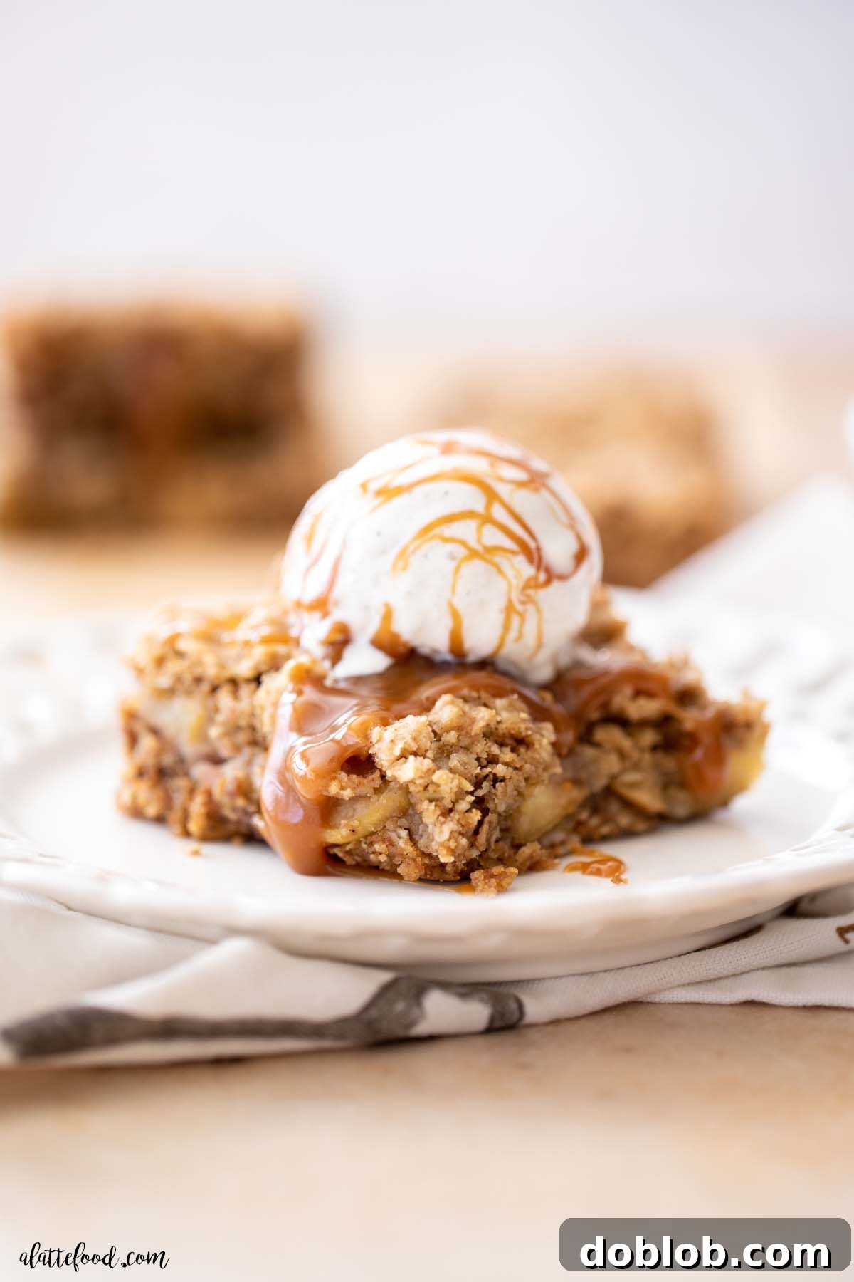 crumb topped apple bar on a white plate with ice cream and extra caramel sauce.