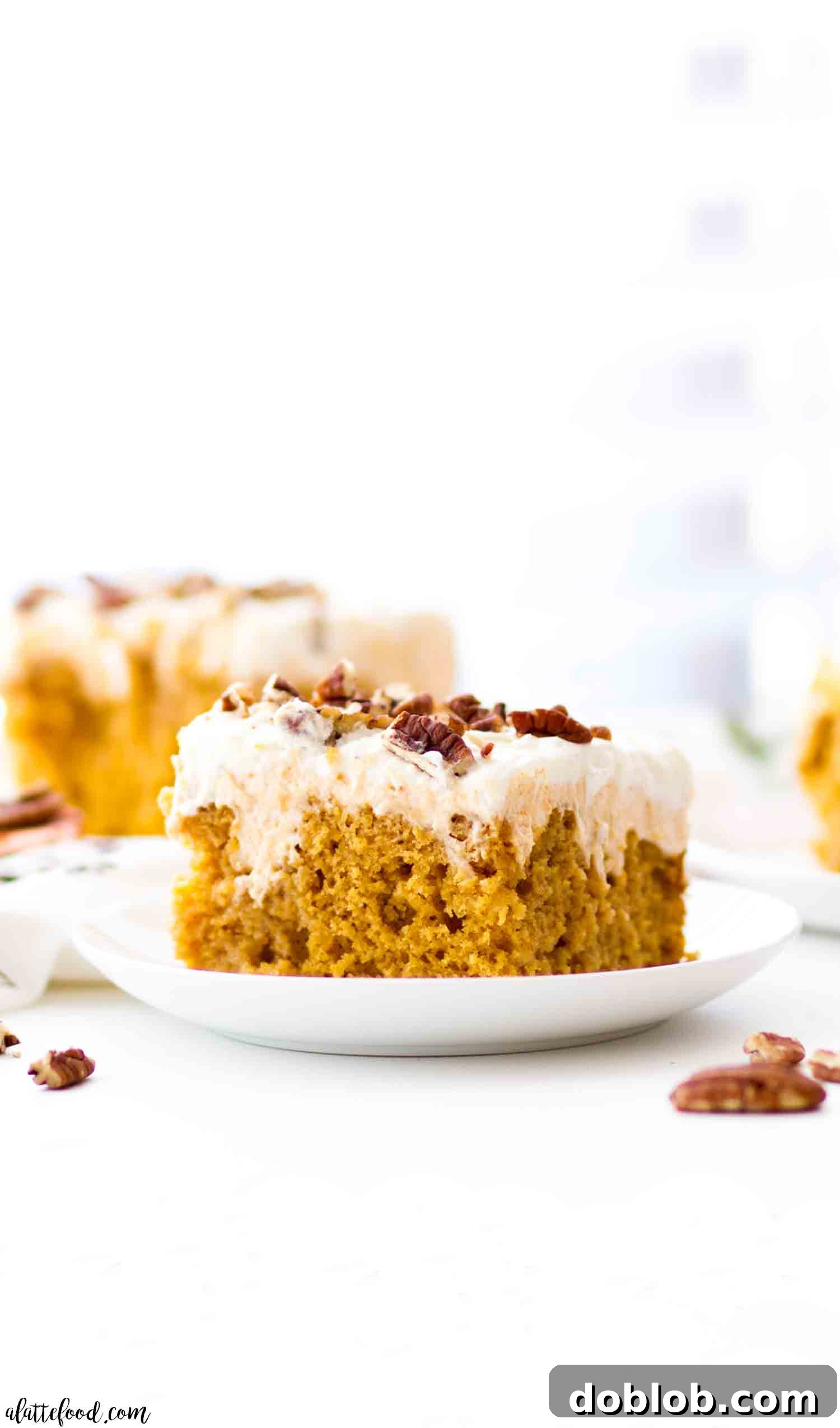 A slice of Pumpkin Poke Cake adorned with whipped cream cheese frosting on a pristine white plate, with the rest of the cake in a pan in the background.