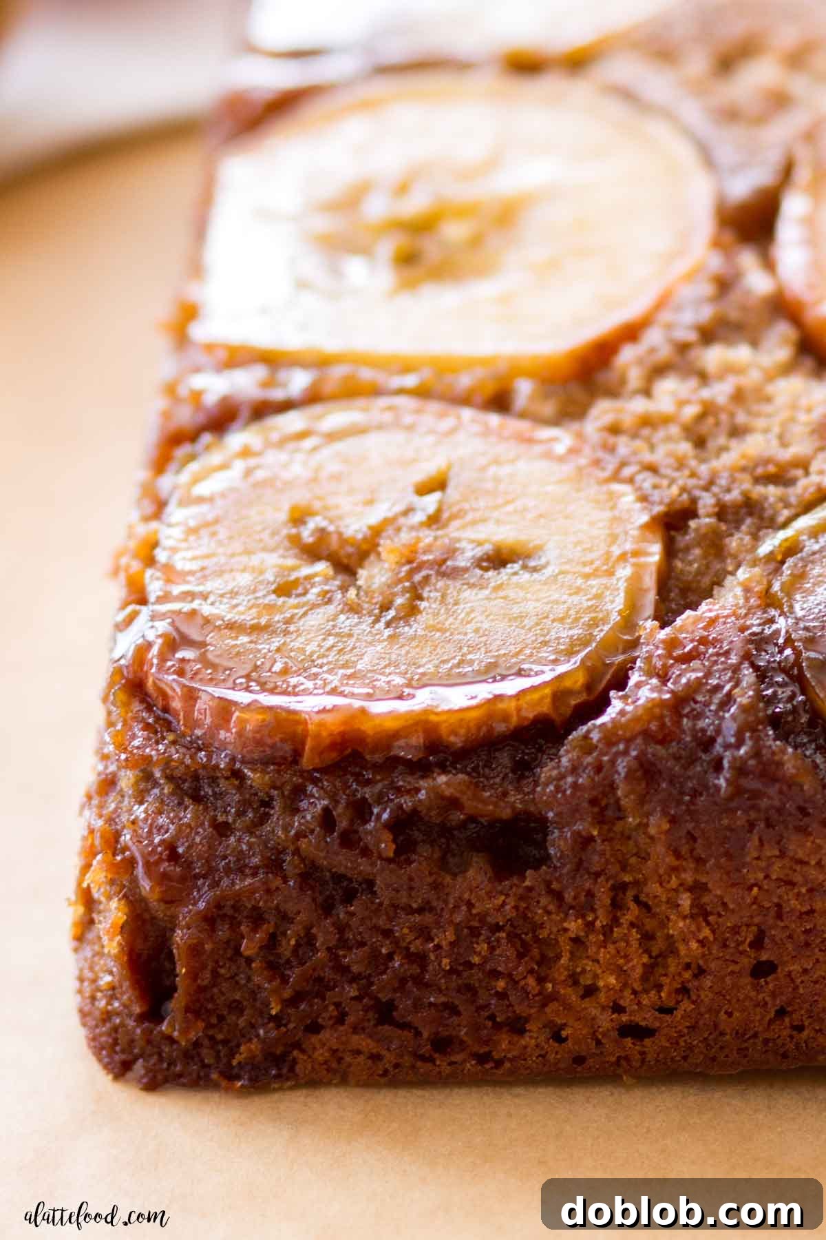A close-up shot of a baked caramel apple cake, still in its square form, resting on brown parchment paper, showcasing the golden-brown crust and caramelized apple topping.