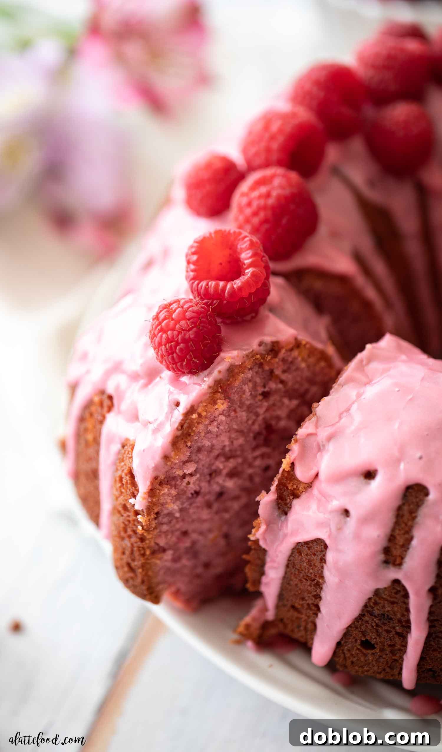 An easy Raspberry Bundt Cake recipe crafted with fresh and freeze-dried raspberries. This from-scratch cake, made without gelatin or cake mix, boasts a light and fluffy texture thanks to simple ingredients like sour cream and milk. Overhead perspective of a perfectly sliced raspberry bundt cake, revealing its delicate pink crumb and a drizzle of glaze, served on a white plate with fresh berries.