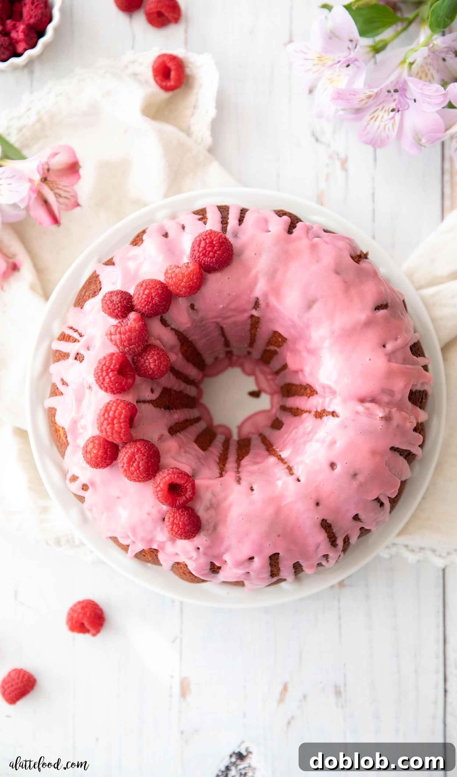 This homemade Raspberry Bundt Cake, made from scratch without gelatin or cake mix, is light, fluffy, and loaded with raspberry flavor thanks to freeze-dried raspberries and a quick raspberry sauce. It’s a beautiful dessert for Valentine’s Day and enjoyable all year long. An exquisite raspberry glazed bundt cake artfully arranged on a white plate, complemented by fresh flowers and a scattering of vibrant raspberries.