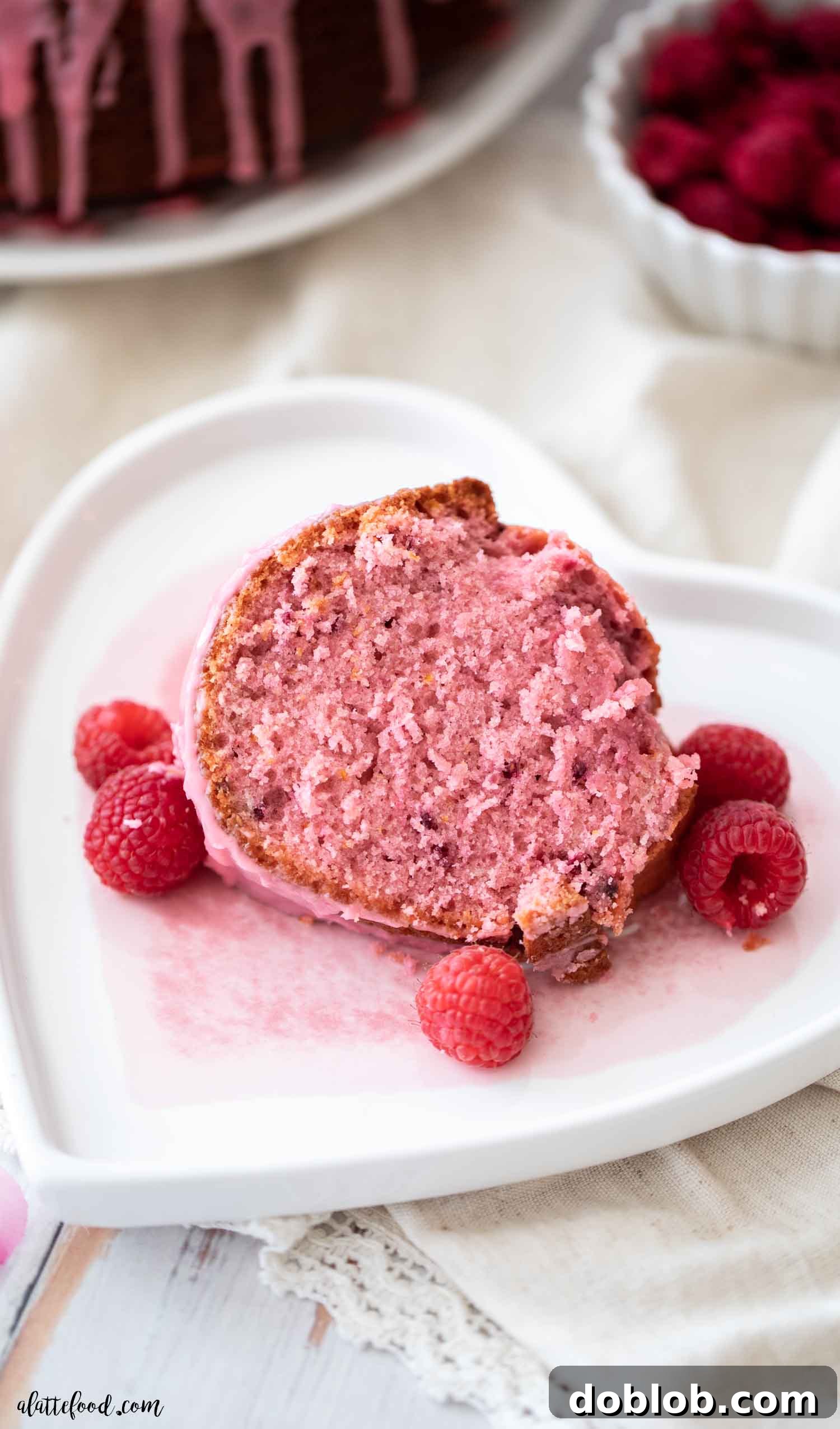 This homemade Raspberry Bundt Cake, featuring a fresh raspberry glaze, is made with fresh and freeze-dried raspberries entirely from scratch – no raspberry jell-o, gelatin, or cake mix! It's an easy baking recipe perfect for spring and Valentine’s Day. A delectable slice of raspberry cake resting on a heart-shaped plate, adorned with fresh raspberries, hinting at a romantic treat.