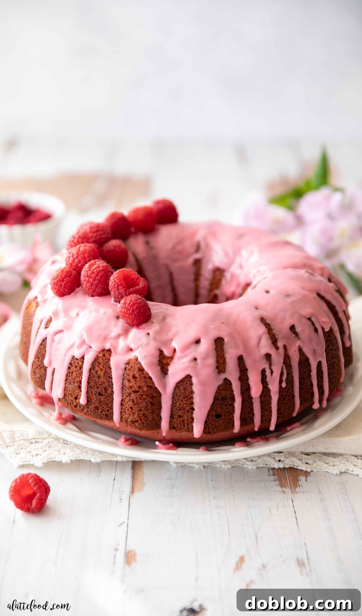 This homemade Raspberry Bundt Cake with a raspberry glaze is made from scratch (a raspberry cake without gelatin or cake mix) using fresh and freeze-dried raspberries. It’s a light and fluffy sour cream bundt cake, close to a raspberry pound cake, and a favorite easy baking recipe for Valentine’s Day. A stunning raspberry glazed raspberry cake presented on a rustic wooden board, adorned with delicate lavender flowers for an elegant touch.