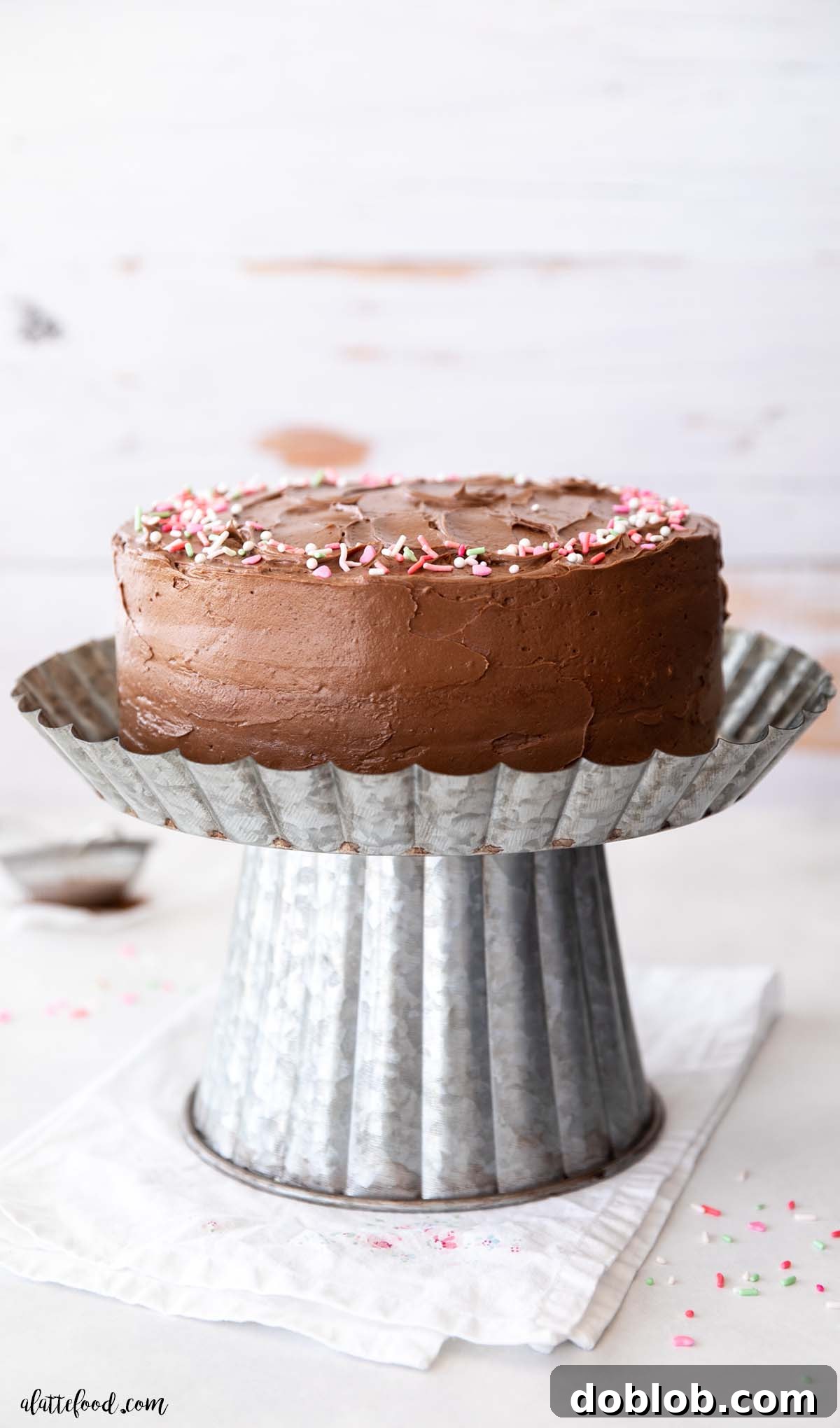 A beautifully frosted yellow cake on an elegant tin cake stand, showcasing its creamy chocolate buttercream and classic appeal.