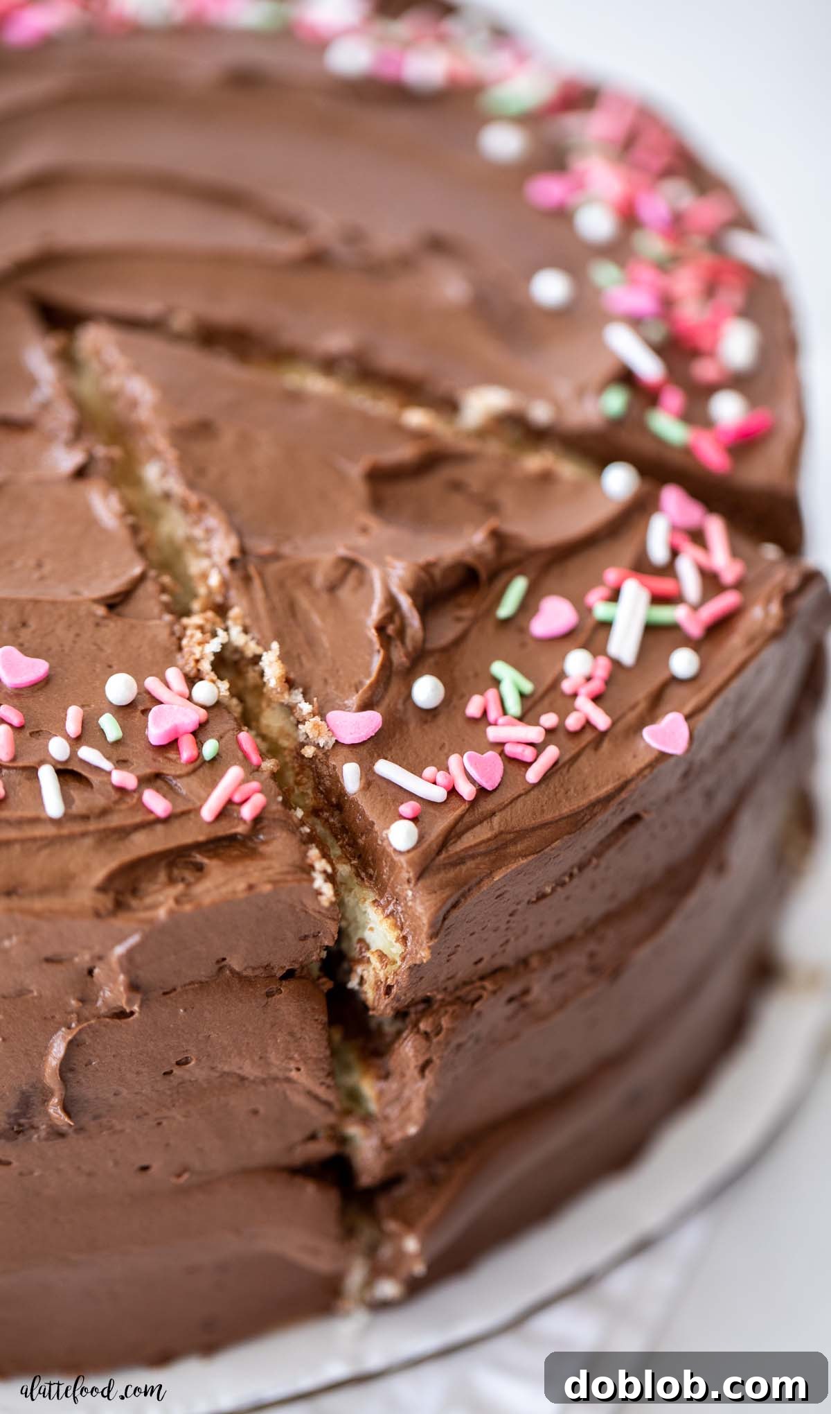 A classic yellow cake, beautifully frosted with chocolate buttercream and adorned with sprinkles, presented on a pristine white cake stand.