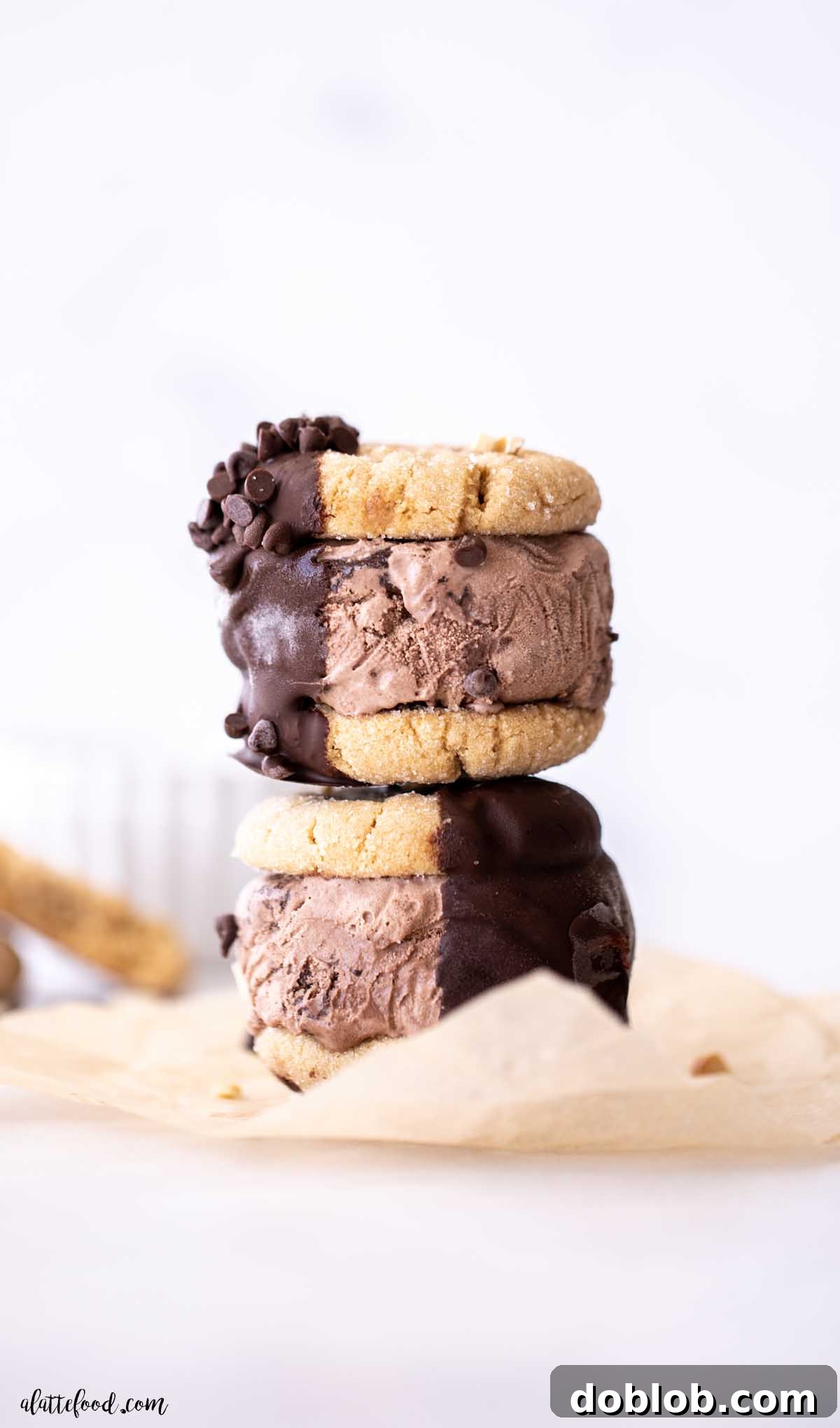 Two peanut butter cookie ice cream sandwiches dipped in chocolate, stacked on parchment paper