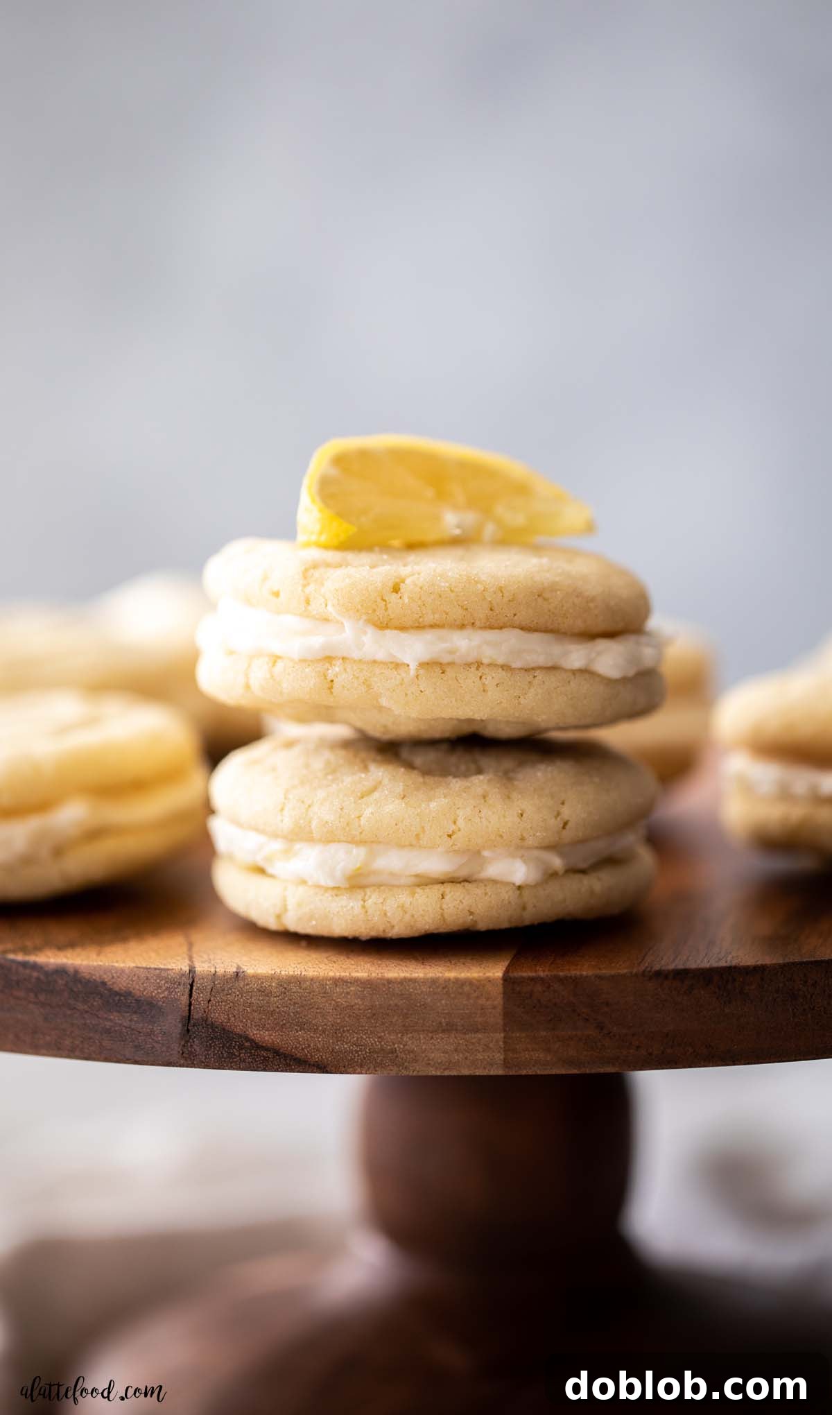 two lemon sugar cookies with lemon buttercream frosting stacked on a wooden cake stand with a lemon wedge on top