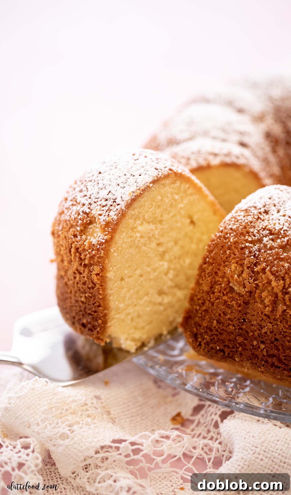 A perfectly golden-brown slice of cream cheese pound cake with a dusting of powdered sugar, served on a elegant silver cake server. The cake exhibits a dense yet tender crumb and moist texture.