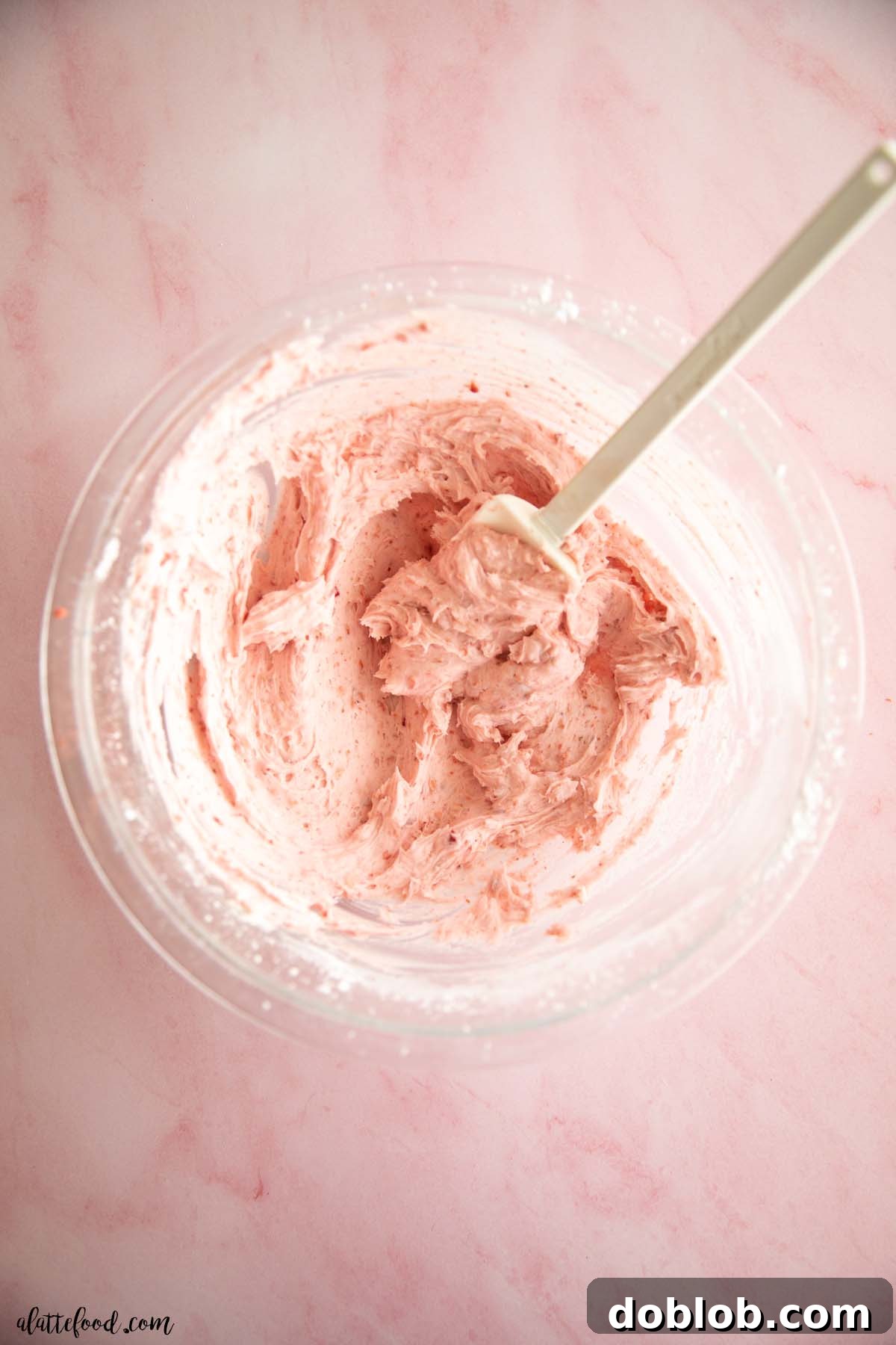 Smooth strawberry buttercream in a glass bowl with a rubber spatula, ready for use.