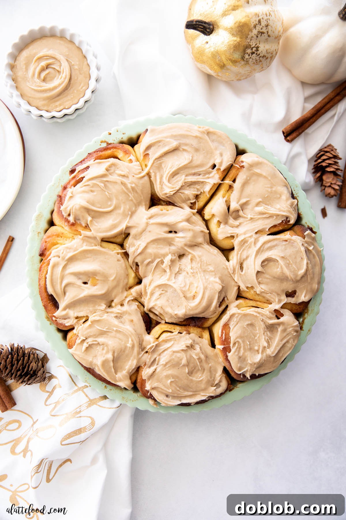 Soft, gooey Pumpkin Butter Cinnamon Rolls with creamy brown sugar frosting, perfect for Thanksgiving brunch. Brown sugar frosted pumpkin cinnamon rolls in a sage pie plate on a white baking board.