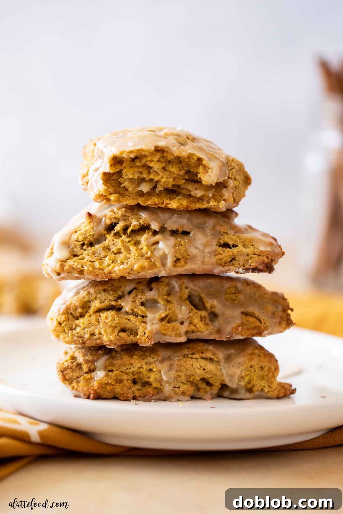 These Buttermilk Pumpkin Scones are wonderfully flaky and tender, crowned with a sweet maple glaze. A stack of golden-brown pumpkin scones, generously drizzled with maple glaze, resting on a elegant white and gold plate.