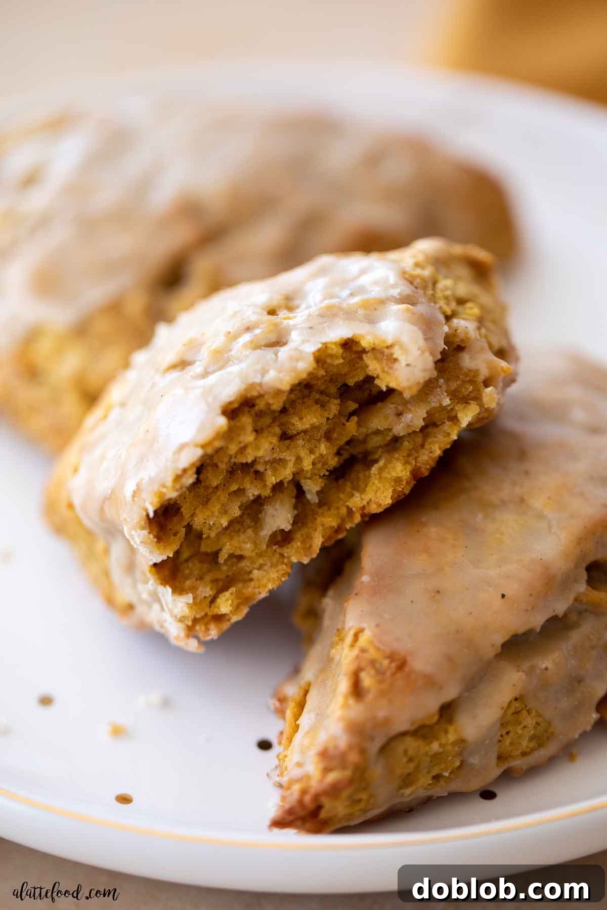 These homemade pumpkin scones are light, flaky, and incomparably topped with a rich maple glaze. A perfectly baked pumpkin scone, sliced in half, revealing its tender, flaky interior, served on a white plate.