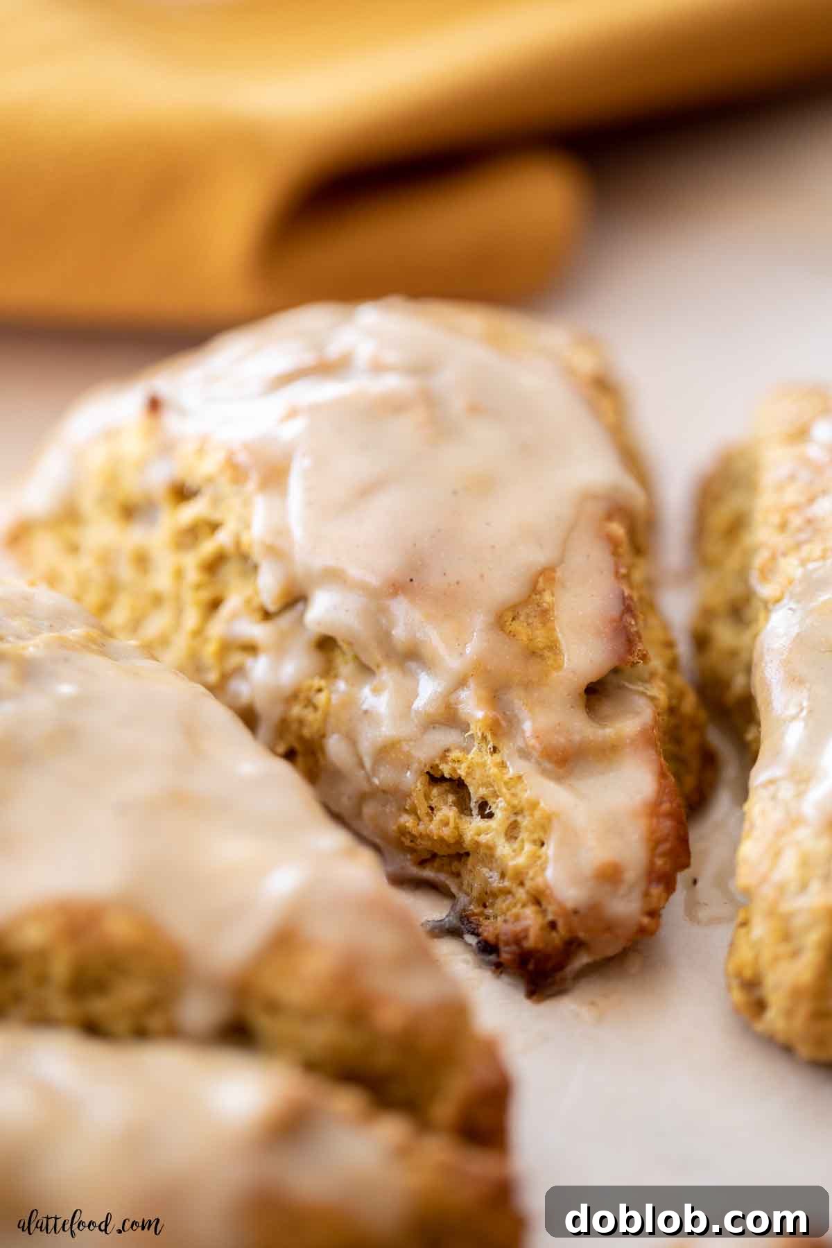 Flaky, buttery, and incredibly easy to make, these maple glazed pumpkin scones are a dream come true. Perfectly baked maple glazed pumpkin scones arranged on parchment paper, with a golden tea towel draped behind them.