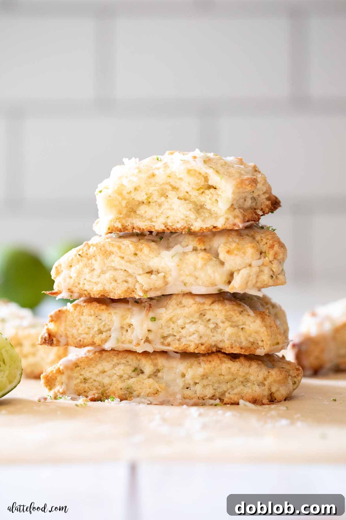 A charming stack of freshly baked coconut lime scones, adorned with a vibrant lime glaze, nestled on brown parchment paper alongside toasted coconut and fresh lime wedges.