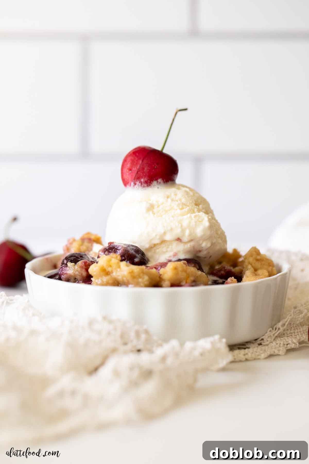 This easy homemade cherry crumble is perfect for summer, featuring fresh or frozen cherries and a crunchy sugar cookie crumb topping with ice cream. Ice cream topped cherry crumble in a white ramekin on a lace towel, showcasing the inviting texture.