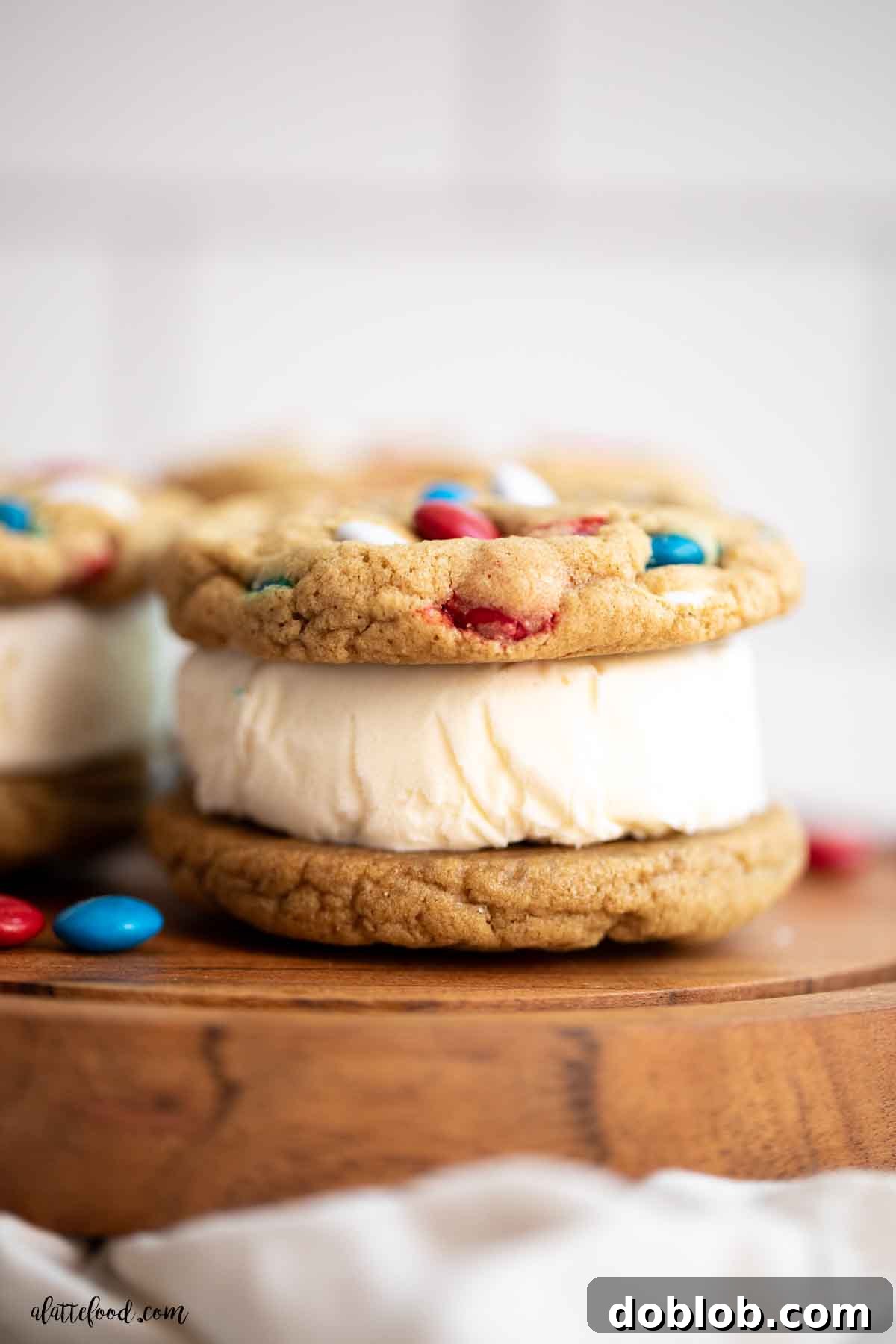 Vibrant M&M cookie ice cream sandwich, featuring homemade soft M&M cookies and rich vanilla ice cream, presented beautifully on a rustic wood cake stand. The perfect summer dessert.