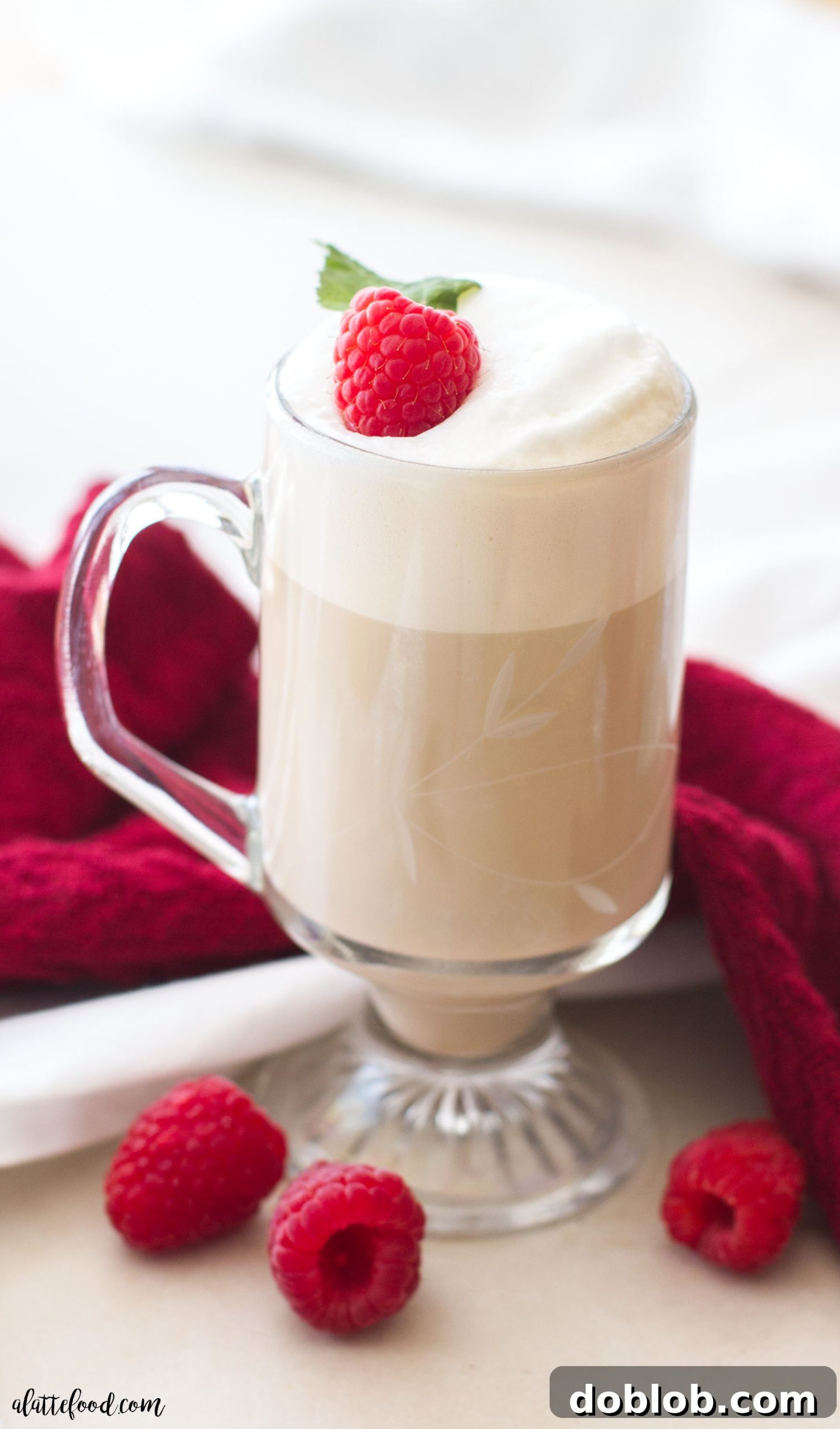 A beautifully crafted White Chocolate Raspberry Vanilla Latte in a clear glass mug, topped with frothed milk and a fresh raspberry.